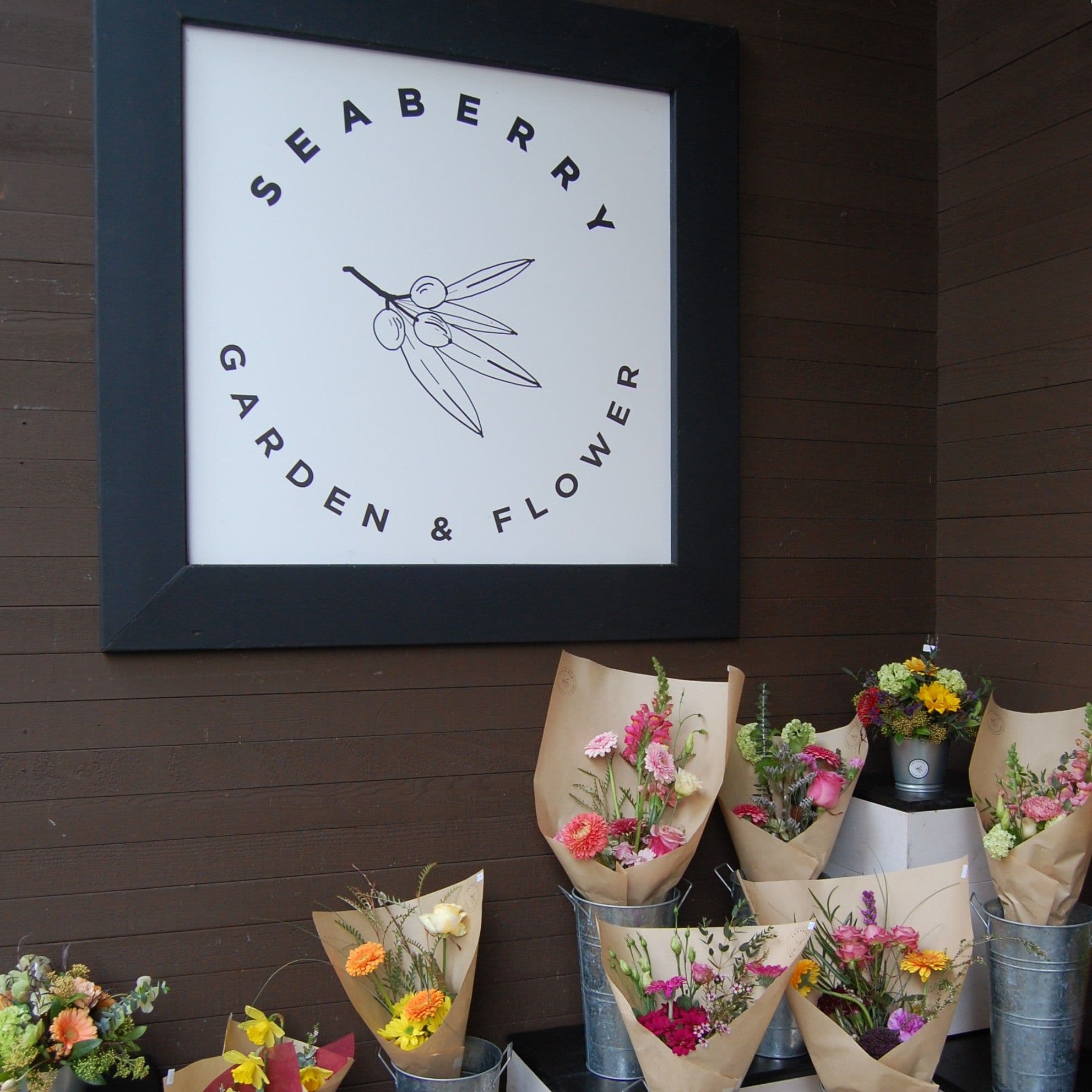 Black-framed Seaberry logo on dark wood beside arrangements in galvanised pails