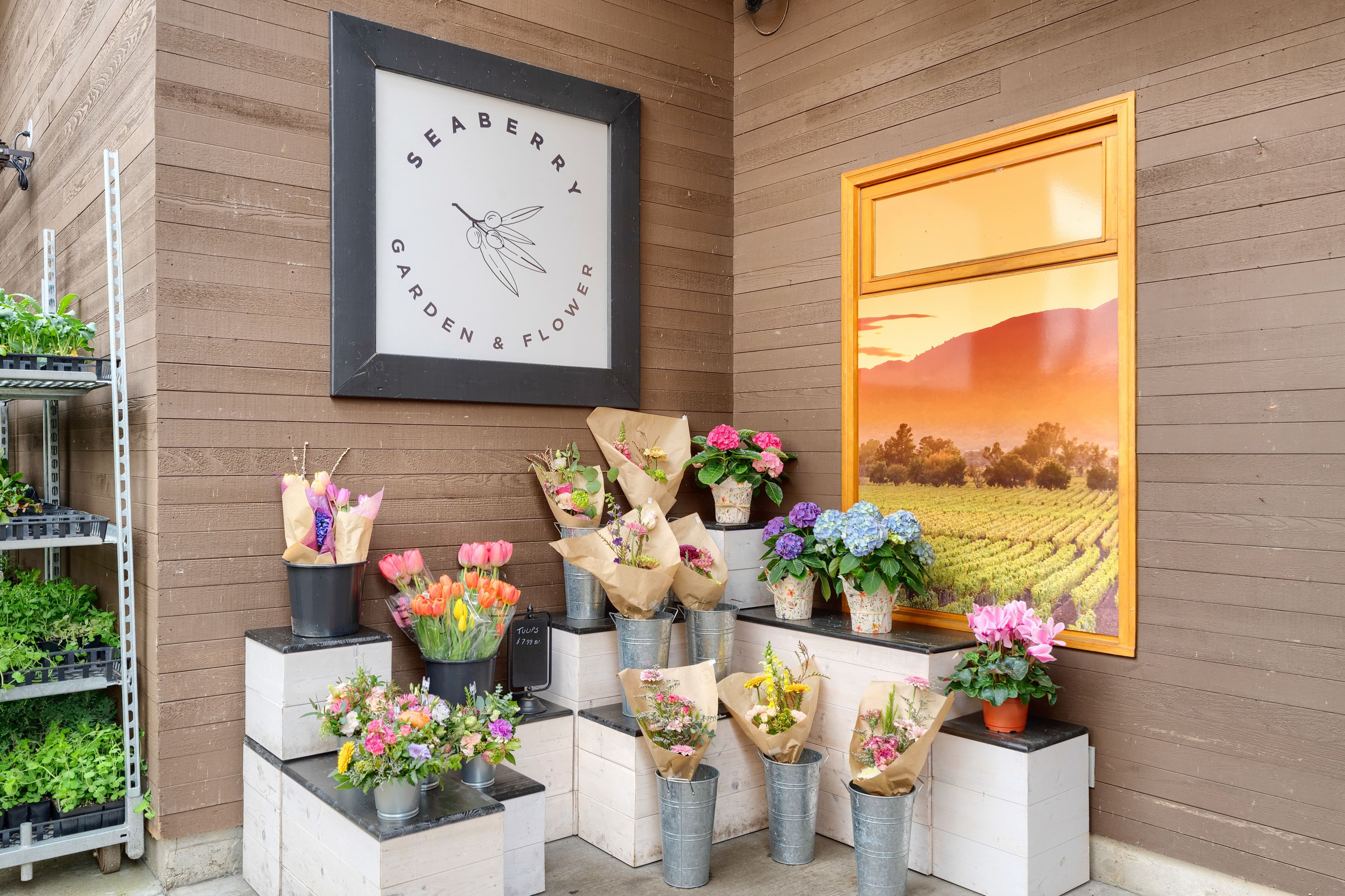 Framed Seaberry Garden & Flower logo above bouquets in tin pails with a sunset landscape painting